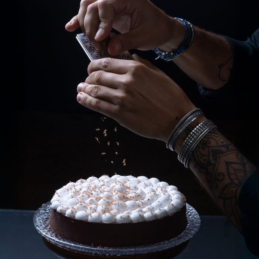 Hands grating over a dark chocolate cake, the falling shavings caught mid-air