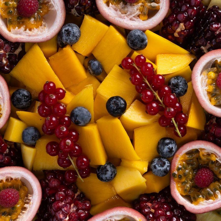 A dense overhead flat lay of mango, berries, passion fruit and pomegranate filling the whole frame