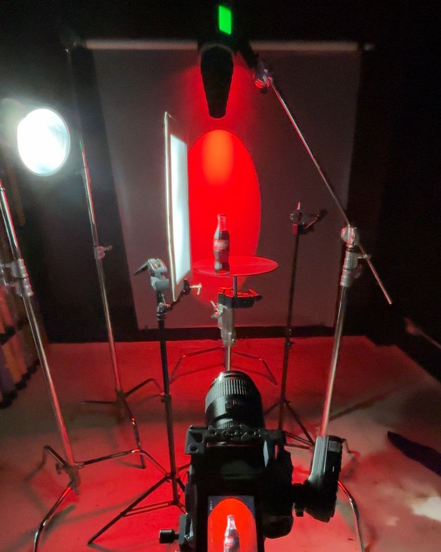 Behind the scenes on a studio shoot — a strip box, boom arms and C-stands surrounding a small red set, with a tethered camera on a tripod